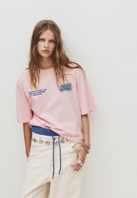 Lyserød bomulds t-shirt med korte ærmer, prydet med blå og hvid trykt tekst og et rektangulært grafik. Bæres med lyse, bælteholdte bukser.
