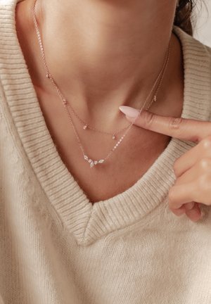 Roségoldene, geschichtete Halsketten mit klaren Edelsteinen, die mit zarten Ketten und verschiedenen Steinformen versehen sind, vor einem sanften cremefarbenen Pullover.