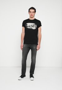 Μαύρο γραφικό t-shirt με την εκτύπωση "DIESEL", συνδυασμένο με στενά γκρι σκούρα τζιν και μαύρα αθλητικά παπούτσια με λευκές λαστιχένιες σόλες.