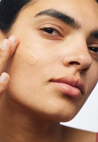 Gros plan sur un visage avec une peau légèrement tachetée de rousseur, appliquant un produit clair en gel, mettant en valeur la texture lisse et la couleur naturelle des lèvres.