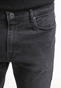 Jean en denim noir présentant un design classique à cinq poches, une fermeture avant à boutons et des détails subtils de couture sur le tissu.