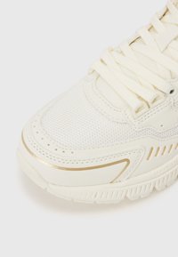 White athletic shoe featuring a textured mesh upper, smooth synthetic overlays, gold accents, and flat laces. Notable logo on the side.