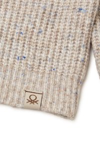 Beige Strickpullover mit blauen Sprenkeln, mit geripptem Muster und einem kleinen Etikett mit einem Logo am unteren Saum.