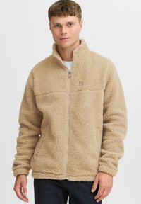 Chaqueta de forro polar beige con cuello alto, cierre de cremallera y superficie texturizada. Presenta un logo en el pecho y un ajuste ligeramente entallado.