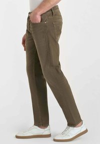 Jean en coton vert olive à coupe slim, avec poches avant et patte de boutonnage. Porté avec des sneakers blanches et un revers de cheville visible.