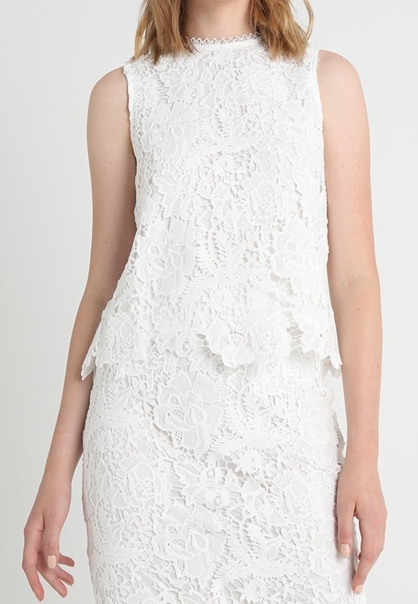 Vitt spetstopp med scallopkant, prydd med blommönster och ärmlös design. Halsringningen är detaljerad med en fin kant.