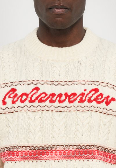 Suéter de punto crema con una textura trenzada, un logo rojo de "Rolandewiler" y rayas horizontales rojas y marrones en el pecho.