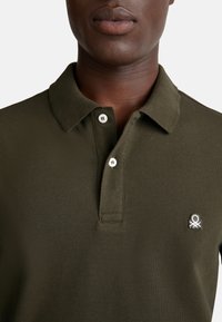 Detailní záběr osoby v olivově zeleném polo tričku se dvěma bílými knoflíky a malým bílým vyšitým logem na hrudi.