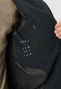 Interno di giacca nera con una tasca con zip e testo stampato "IKKS URBAN LAB." Presenta un mix di tessuto e materiale liscio.