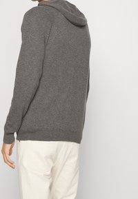 Grauer gestrickter Kapuzenpullover mit weicher Textur, ausgestattet mit einer Kordelzugkapuze und gerippten Bündchen, kombiniert mit hellen Hosen.