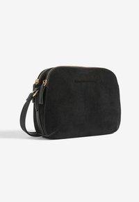 Borsa crossbody in suede nero con doppia cerniera e tracolla regolabile, con testo inciso in modo sottile sulla parte anteriore, su uno sfondo bianco semplice.
