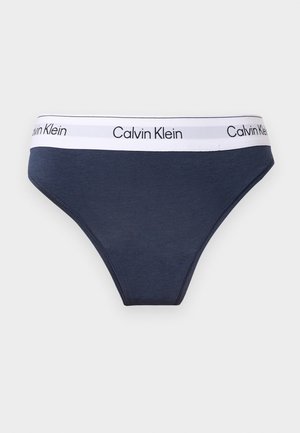 Slip Calvin Klein bleu marine avec une ceinture élastique blanche comportant le nom de la marque en texte noir, présenté sur un fond gris clair.