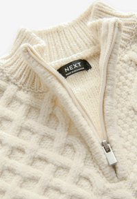 Pull en maille couleur crème avec motif géométrique, demi-fermeture éclair et étiquette noire NEXT indiquant « Made in Myanmar ».