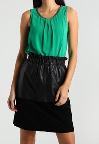 Haut sans manches vert avec des plis et une bordure brillante, associé à une taille en cuir noir et une jupe noire au toucher doux.