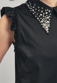 Top senza maniche nero con fori per le braccia arricciati e un colletto statement decorato con perle e strass, che presenta una texture liscia.