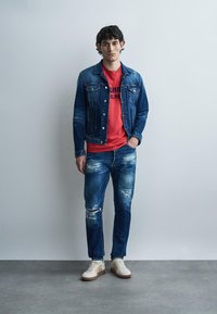 Fiatal férfi világos fal előtt áll, kék farmer dzsekit, kopott kék farmert, piros mintás pólót és fehér sportcipőt visel.