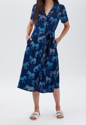 Woman wearing a navy blue dress with light blue zebra prints, short sleeves, button front, waist tie, and white sandals with buckles.