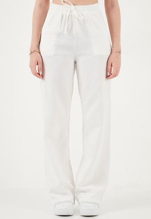 Žena oblečená v voľne padavých bielych nohaviciach so šnúrkou v páse a bielych teniskách, stojaca pred jednoliatym bielym pozadím.