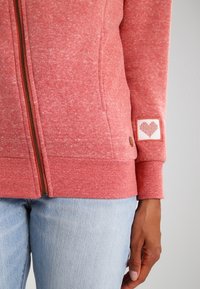 Sudadera con cremallera en coral desvaído, con acabado texturizado, puños acanalados y parche de corazón en la manga. Detalle de cremallera en marrón.