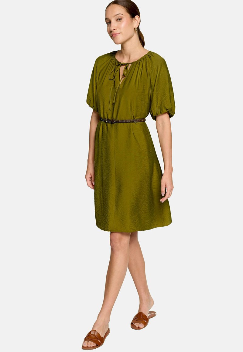 Femme portant une robe vert olive longueur genou avec des manches bouffantes et une ceinture marron, associée à des sandales plates marron, debout sur fond blanc.