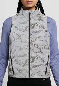 Gilet senza maniche grigio chiaro con motivi astratti neri e metallici, dotato di un colletto alto, cerniera nera e tasche laterali con cerniera.