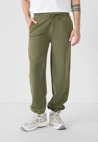 Olijfgroene sweatpants met een trekkoord in de taille, zijzakken, taps toelopende pijpen en een zachte textuur, gecombineerd met lichtgrijze en witte sneakers.