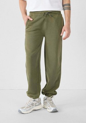 Pantalones deportivos verde oliva con cintura ajustable, bolsillos laterales, perneras ajustadas y textura suave, combinados con zapatillas ligeras en gris y blanco.