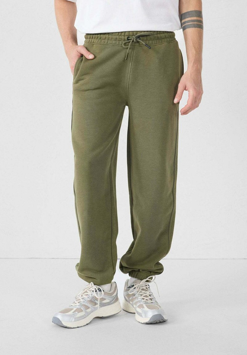 Olijfgroene sweatpants met een trekkoord in de taille, zijzakken, taps toelopende pijpen en een zachte textuur, gecombineerd met lichtgrijze en witte sneakers.