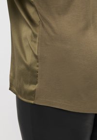 Top verde oliva con una texture liscia e satinata sui lati e una finitura opaca sul corpo principale; presenta un orlo semplice.