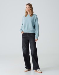 Felpa azzurro chiaro con collo a giro e polsini a costine, abbinata a jeans neri a gamba larga e sneakers bianche con suola marrone.