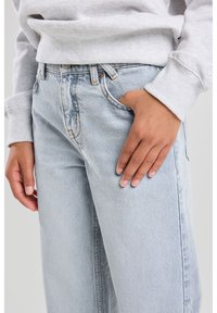 Light blue denim jeans with a relaxed fit, featuring visible stitching, a mid-rise waist, and a hand placed on the thigh area.