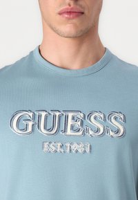 T-shirt in cotone azzurro chiaro con il logo bianco "GUESS" in evidenza e cuciture blu, e "EST. 1981" sottostante. Collo rotondo e maniche corte.
