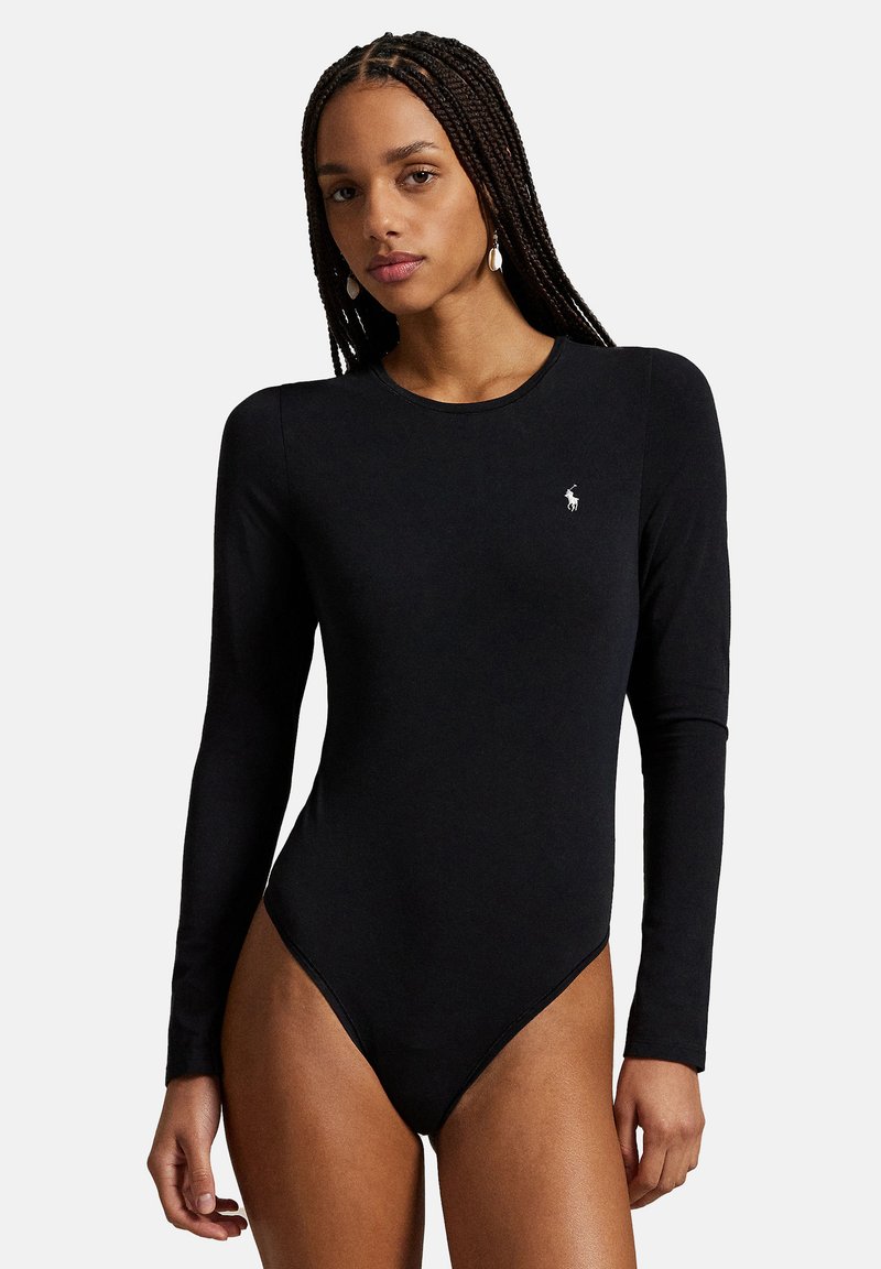 Femme aux cheveux tressés portant un justaucorps noir à manches longues et ajusté avec un petit logo blanc sur la poitrine, debout devant un fond uni.