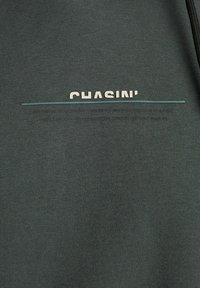 Pullover grigio con tessuto testurizzato, con il testo "CHASIN'" in crema e una linea blu sotto. Dettaglio zip sul lato.