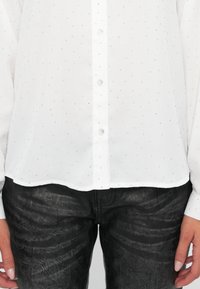 Witte blouse met kleine zilverkleurige stippen, knoopsluiting, lange mouwen en een rechte zoom. Gecombineerd met zwarte structuurpants.