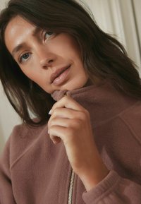 Close-up van een vrouw met lang, donker haar, gekleed in een zachte, mauve fleecejas, die de rits met één hand vasthoudt en dromerig naar de camera kijkt.