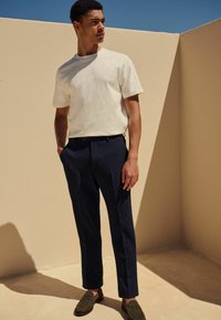 T-shirt blanc à manches courtes associé à un pantalon texturé bleu marine et des mocassins vert foncé, se tenant devant un fond de couleur claire.