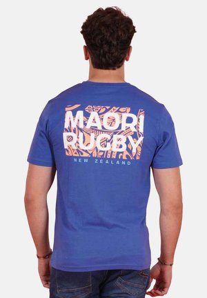 Homme portant un T-shirt bleu avec l'inscription « Māori Rugby New Zealand » et des motifs tribaux orange dans le dos, vu de dos.