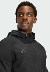 Chaqueta deportiva negra con un cuello alto, cierre de cremallera y detalle de tres rayas en el pecho. Presenta una textura suave y ligera.