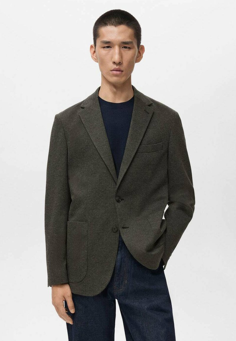 Blazer texture verde scuro con rever, due tasche frontali e chiusura a un bottone, indossato su una camicia blu navy e abbinato a jeans blu.