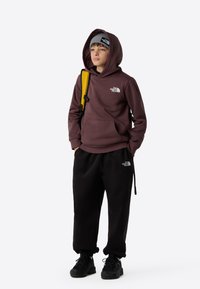 Mørkerød hettegenser med hvit logo, svarte joggebukser, grå lue med logo, og svarte tursko. Inkluderer en gul ryggsekkstropp.