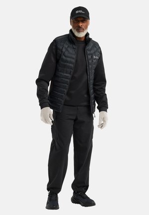 Gilet matelassé noir avec fermeture éclair à l'avant, sweatshirt noir et pantalon cargo. Des gants et des chaussures noirs viennent compléter la tenue. La casquette arbore un logo.