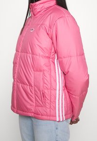 Rosa isolerad jacka med hög krage, sidofickor och vita ränder längs ärmarna. Har en vadderad textur och logotypdetalj.