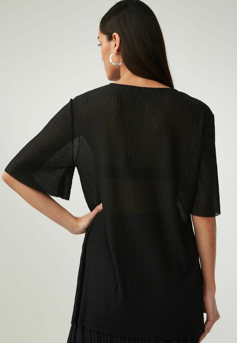 Sort plisseret bluse med korte ærmer, gennemsigtig tekstur og en let forlænget bagkant. Minimalistisk design med en afslappet pasform.