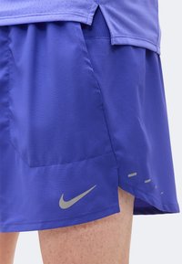 Μωβ αθλητικό σορτς με πλαϊνή τσέπη και μικρό ανακλαστικό λογότυπο Nike κοντά στο τελείωμα, φορεμένα με ένα αντίστοιχο μωβ τοπ.