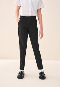 Black tapered trousers made of smooth fabric with a button closure, side pockets, and a plain design, paired with black shoes and white socks.