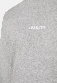 Camisa de algodón gris de manga larga con un pequeño logo blanco "LES DEUX" en la parte superior izquierda del pecho. Textura suave y diseño casual.