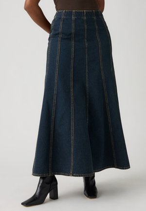 Jupe longue en jean bleu foncé avec des coutures verticales, portée avec des bottines noires à talons et un top marron.