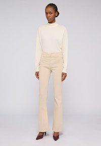 Pantalones beige de tiro alto con diseño de pierna acampanada, combinados con una blusa de manga larga color crema suave, llevados con tacones de aguja color burdeos.