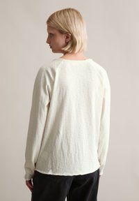 Langärmliger, hellcremefarbener Pullover mit lockerem Schnitt. Weicher, strukturierter Stoff, Rundhalsausschnitt mit ungesäumtem Saum und Raglanärmeln.